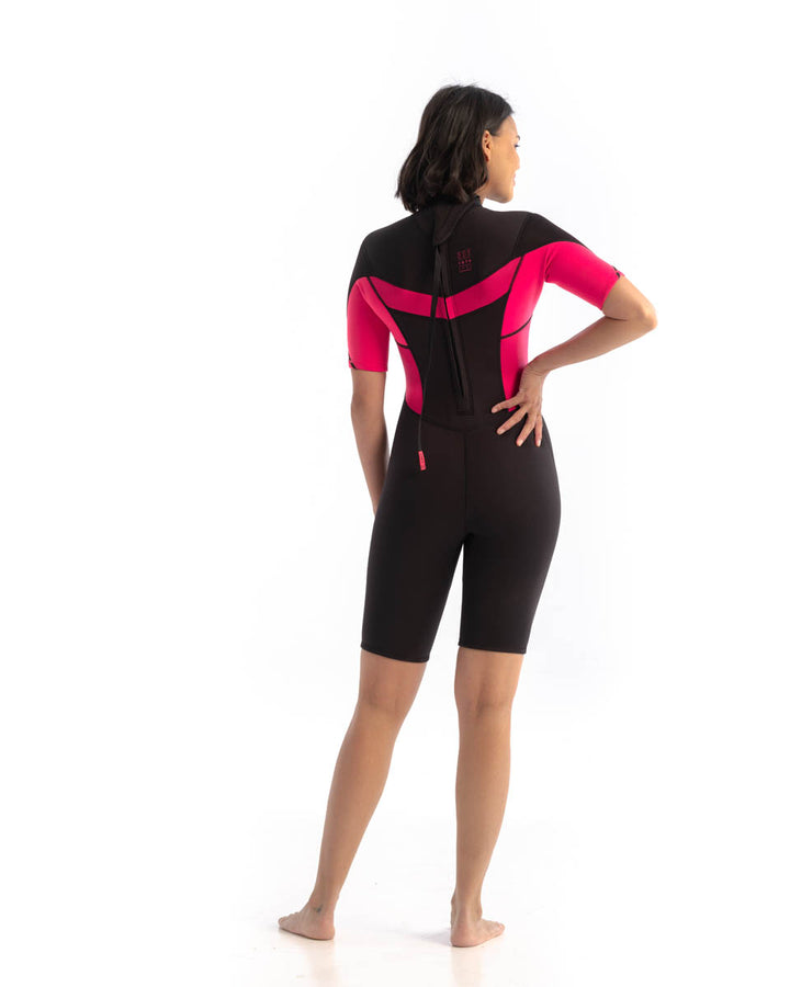 A woman standing, wearing a  black and hot pink Jobe womens shorty wetsuit with short sleeves, short legs and a back zip. View of the back.