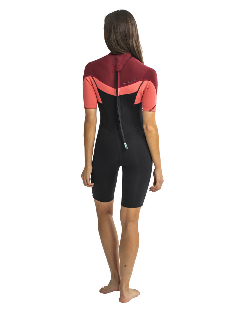 A woman standing, wearing a red, pink and black Jobe womens shorty wetsuit with short sleeves, short legs and a back zip. View of the back.