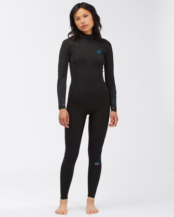 A woman standing and posing in a black full-body Billabong Synery 5/4 womens wetsuit with a back zip entry.