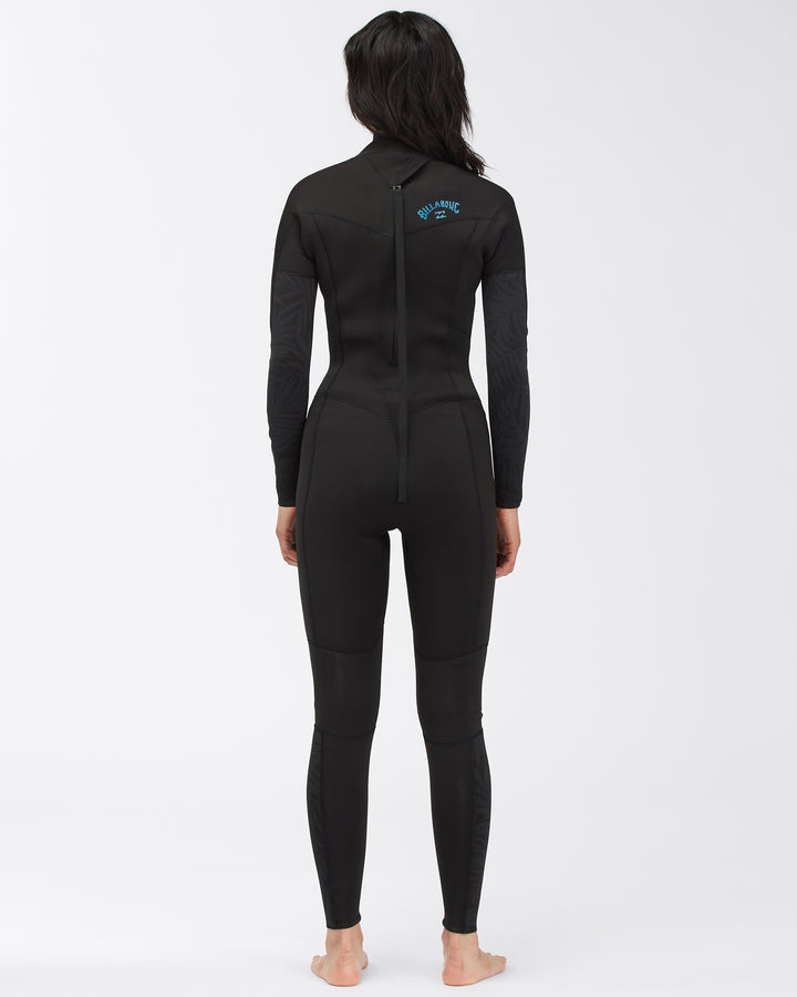 A woman standing with her back facing and posing in a black full-body Billabong Synery 5/4 womens wetsuit with a back zip entry. 